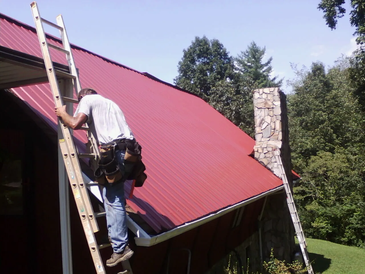 Roofer on a ladder beside a red metal roof and stone chimney — Soffit & Fascia Repair by Roofing Brothers in Terra Alta